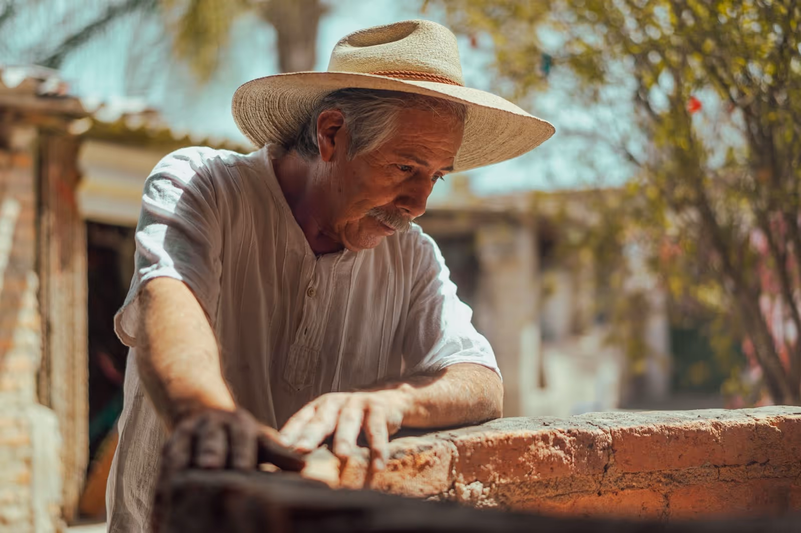 An elderly man with gray hair looks down a well with dirt on his hands. He wears a sun hat to provide shade to his head from a blistering sun