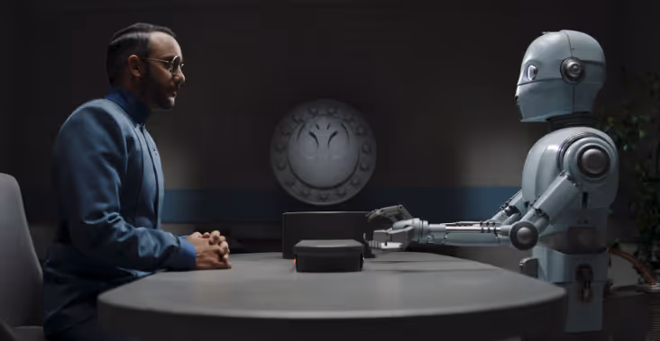 A man wearing a blue uniform sits with his hands together looking across a table in a dark room at a white robot who has its hands out as if speaking