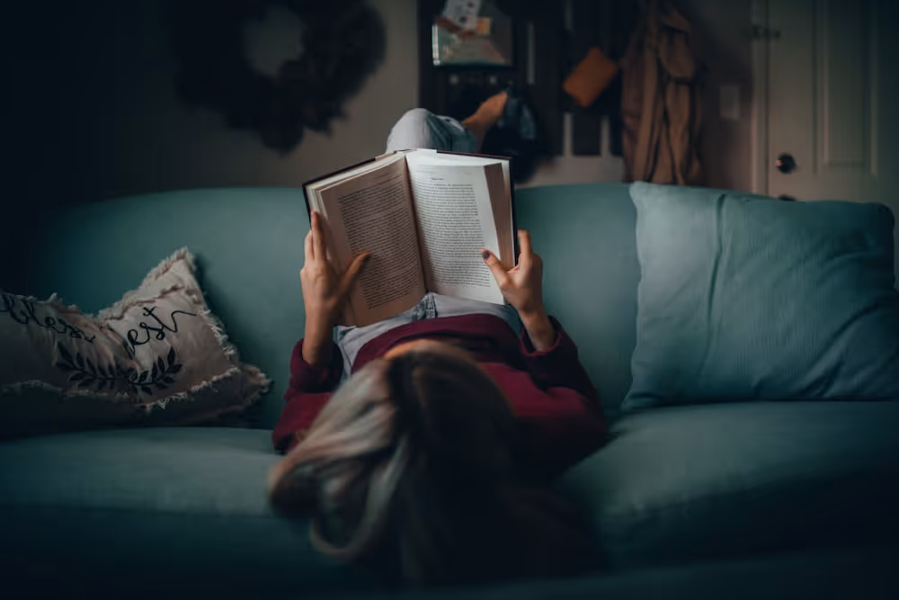 Woman sitting upside down on a couch reading a book. 
