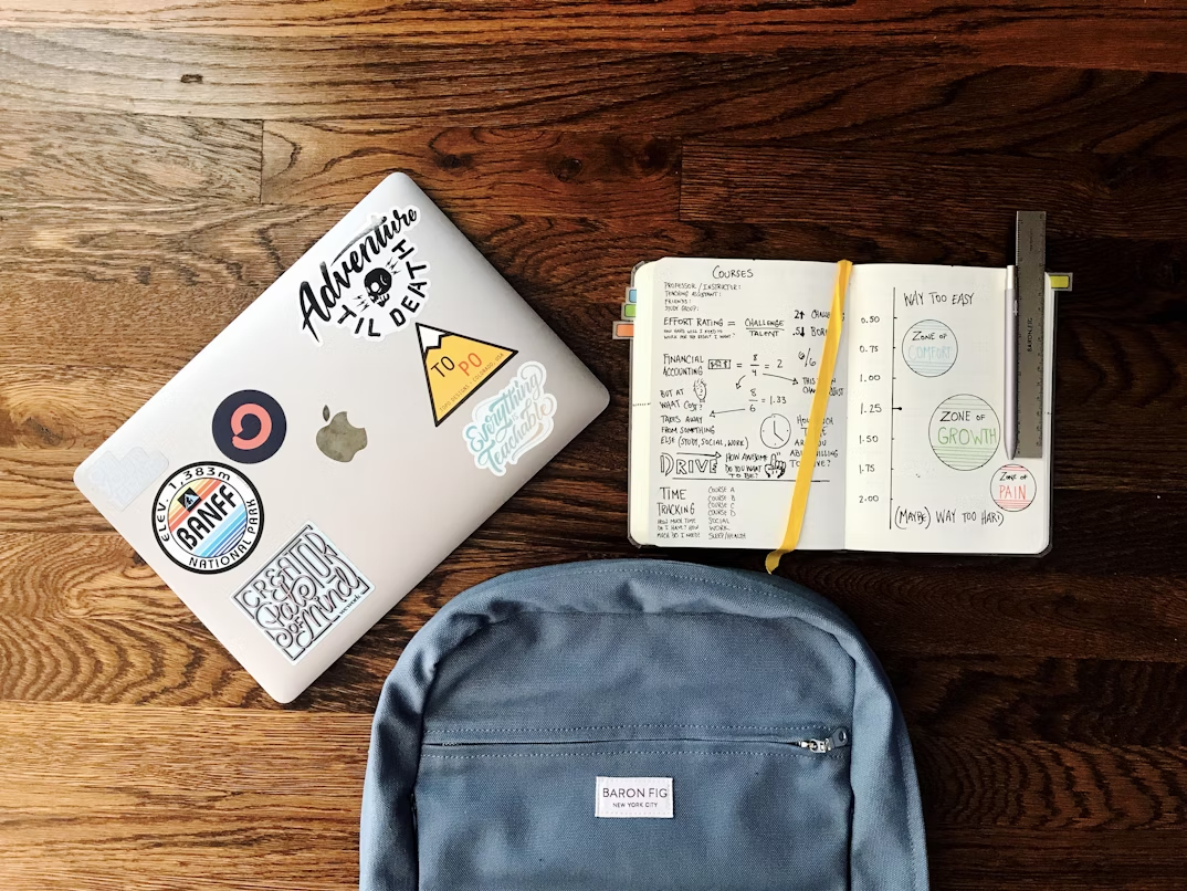 A flat lay photograph of a blue backpack beside a book and a silver MacBook