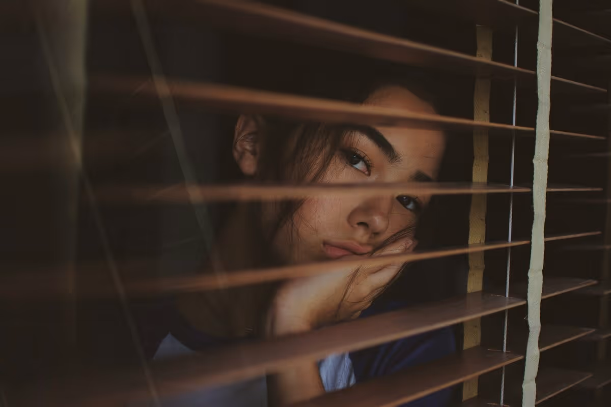 A person looking through opened window blinds. They’re resting their chin in their hand, and seem to be deep in thought.