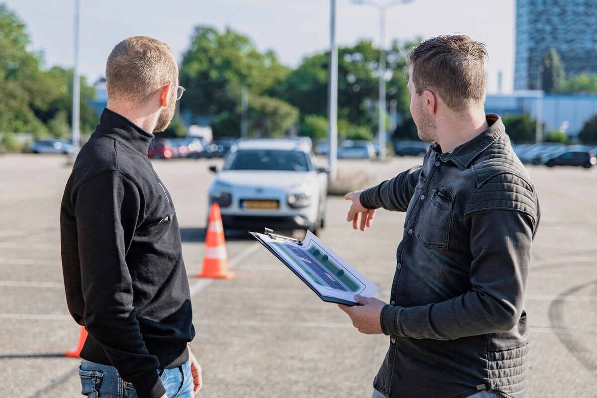 Two people standing in a parking lot. They face away from the camera. One holds a clipboard, and is pointing toward a parked car.