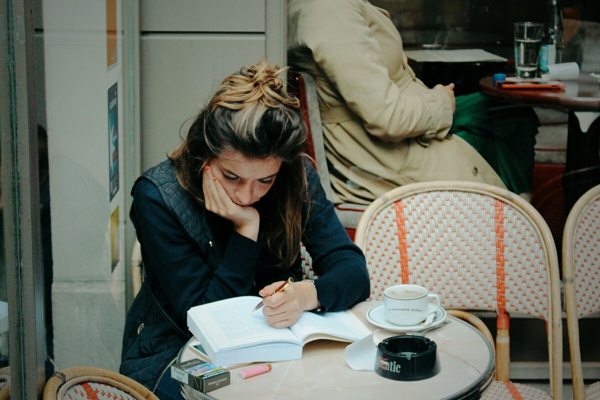 A person with long hair seated in a cafe, focusing on a large book. They appear to be writing in it.