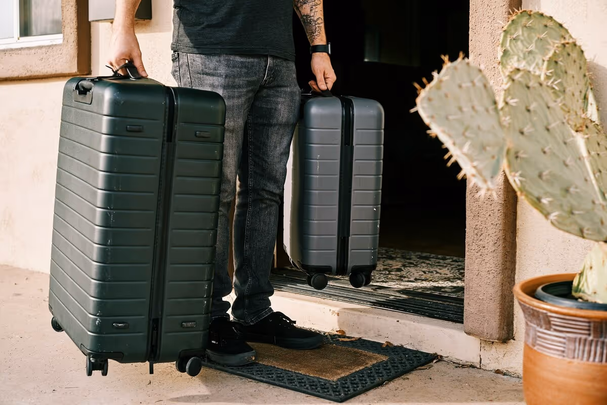 A person holding two suitcases, one large and small. They stand outside a house.