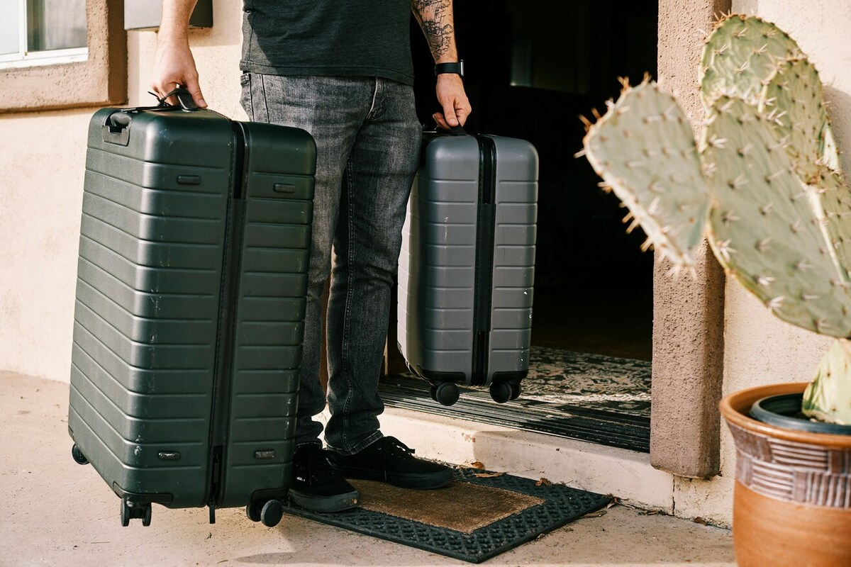 A person holding two suitcases, one large and small. They stand outside a house.