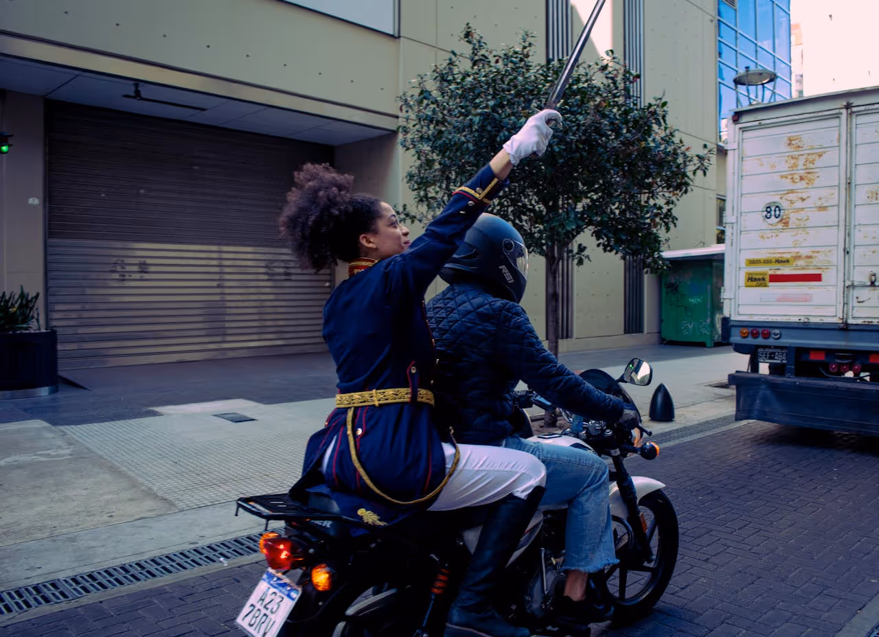couple on a motorcycle with the woman sitting behind the driver raising a sword to call to mind the chivalrous salute of a knight