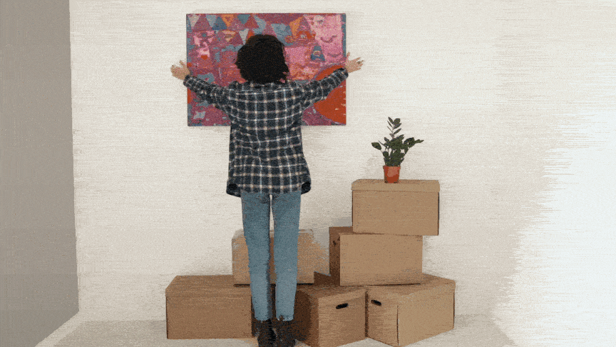 young woman in a flannel shirt hanging a painting on the otherwise blank white walls of her first apartment, moving boxes still piled at her feet