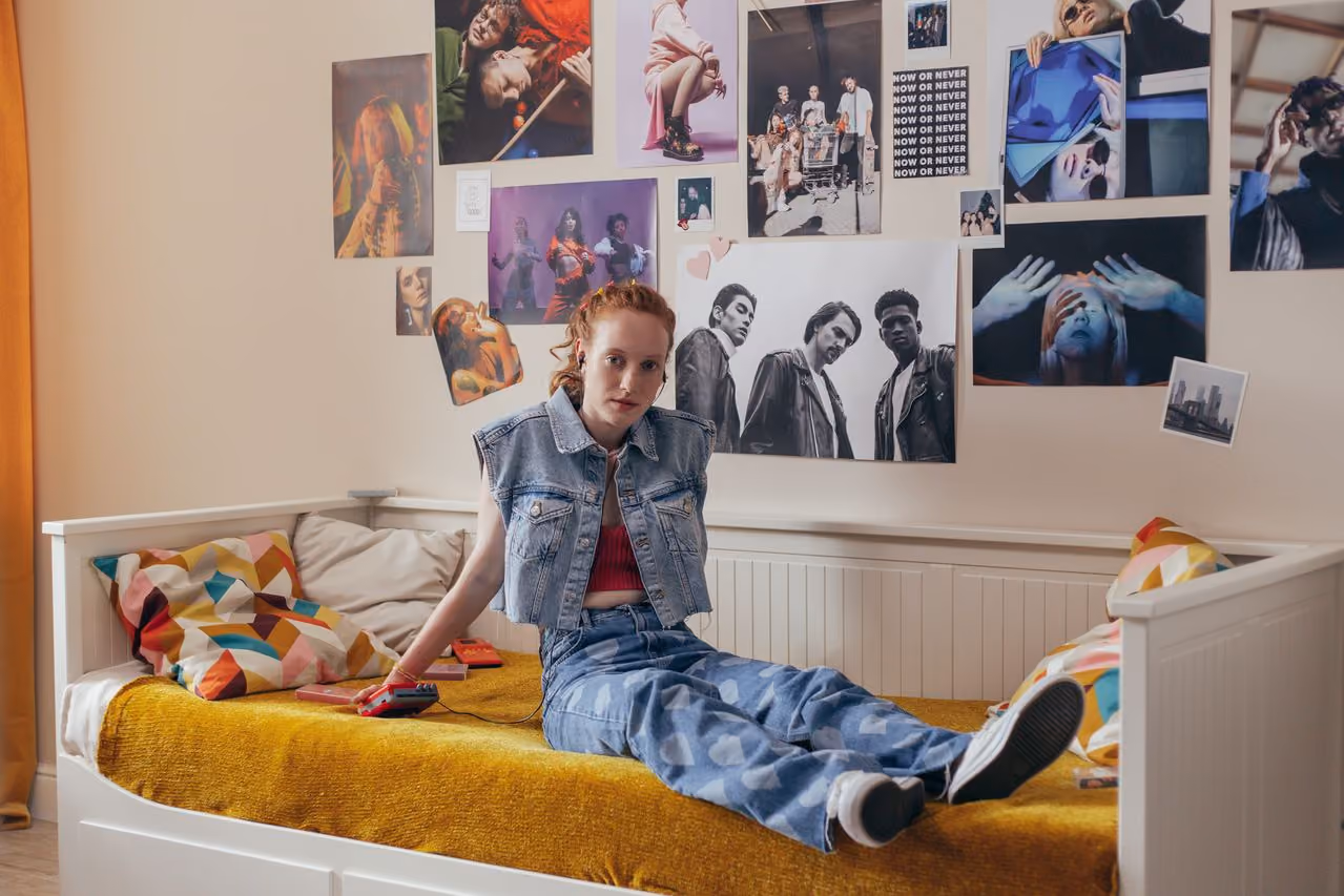 red haired college girl sitting in front of a wall decorated with posters and photos