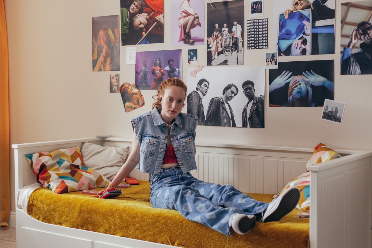 red haired college girl sitting in front of a wall decorated with posters and photos