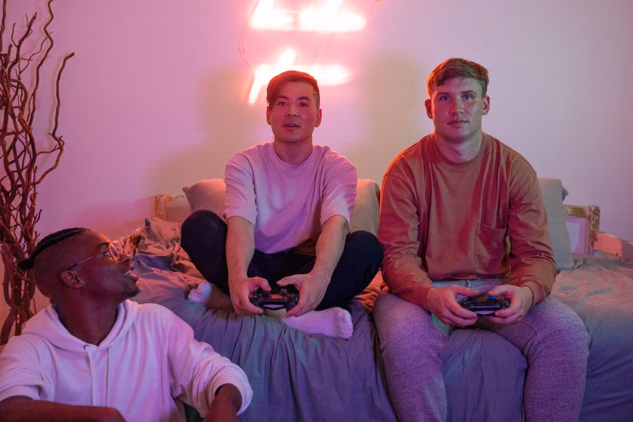 three male college students sitting on a bed with gaming controllers in their hands, lit by the neon glow of their LED light strip