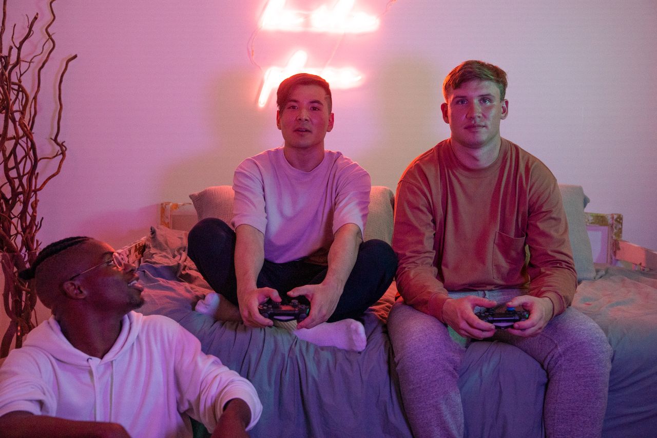 three male college students sitting on a bed with gaming controllers in their hands, lit by the neon glow of their LED light strip