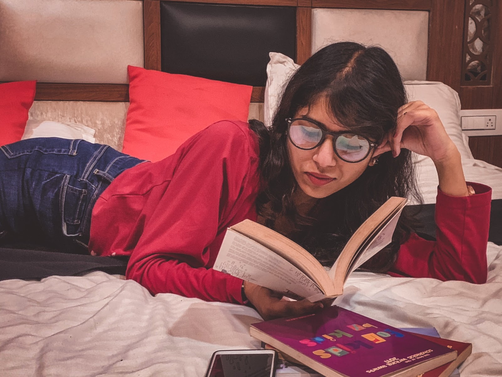 college student lying in bed with a book and several more piled up next to her