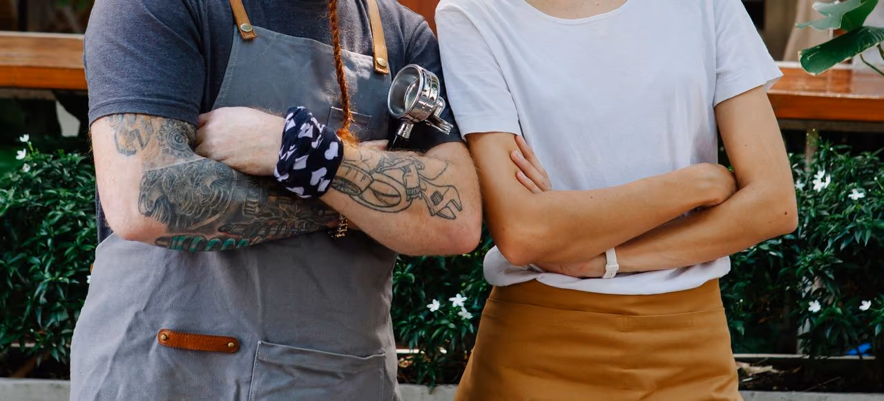 two baristas with their arms folded standing side by side, one with visible tattoos and the other with none, representing the question about whether or not to get a tattoo