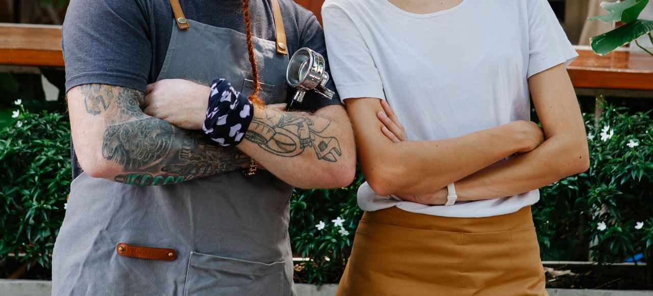two baristas with their arms folded standing side by side, one with visible tattoos and the other with none, representing the question about whether or not to get a tattoo