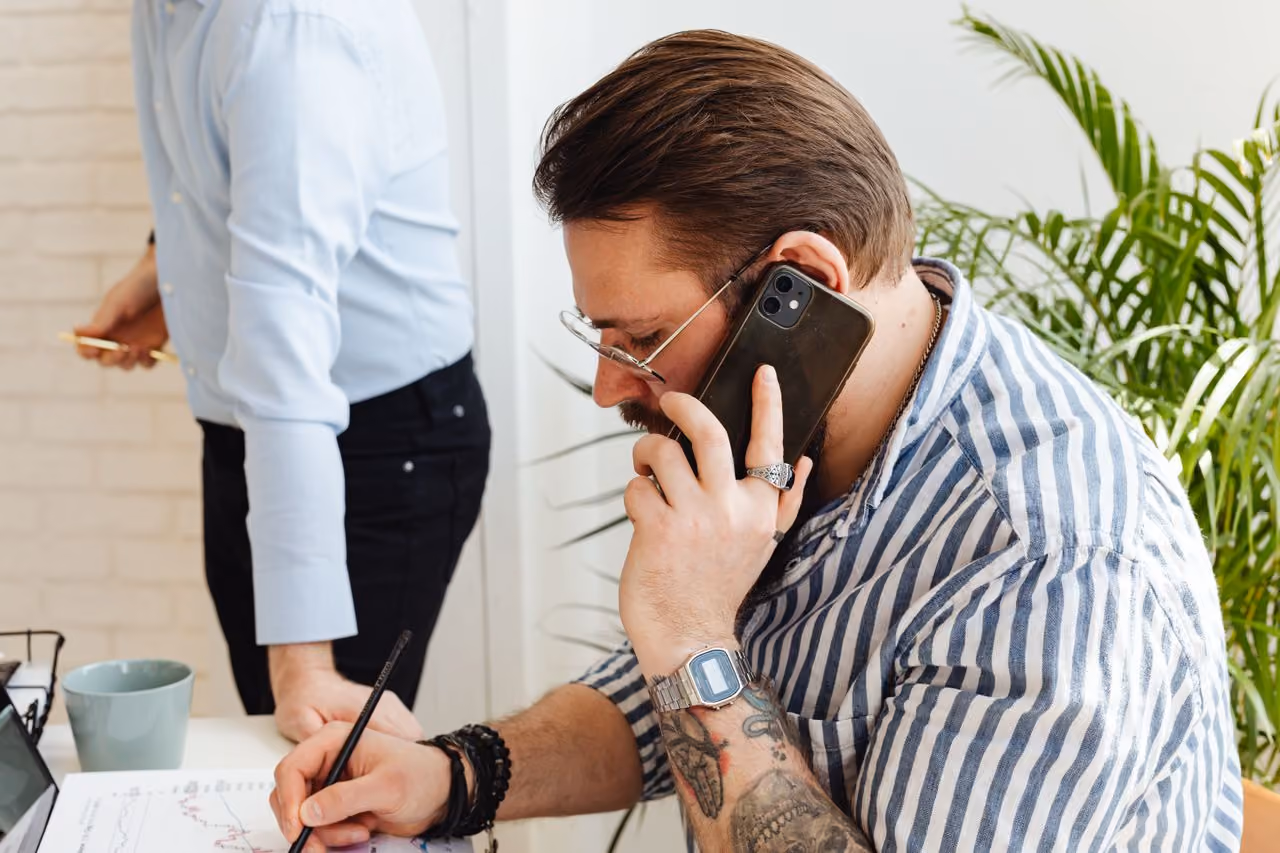 professional adult man wearing a short sleeve button down shirt, speaking on the phone in an office setting, with visible tattoos on his arms