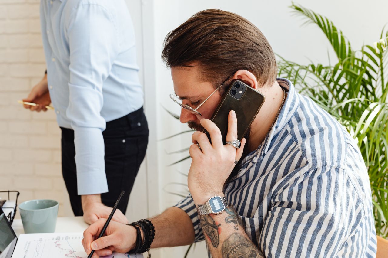 professional adult man wearing a short sleeve button down shirt, speaking on the phone in an office setting, with visible tattoos on his arms