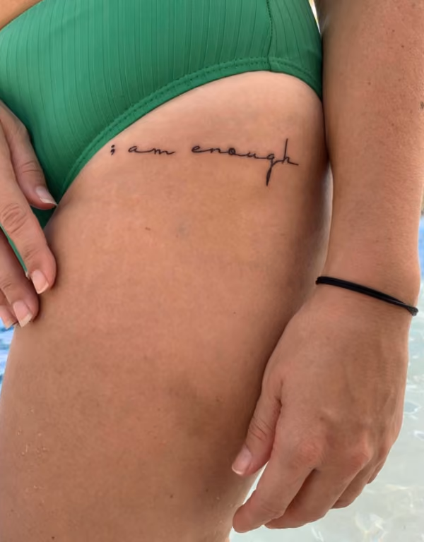 woman in a green swimsuit with I am enough written in script on her upper thigh, with the "I" being a semi colon mental health tattoo