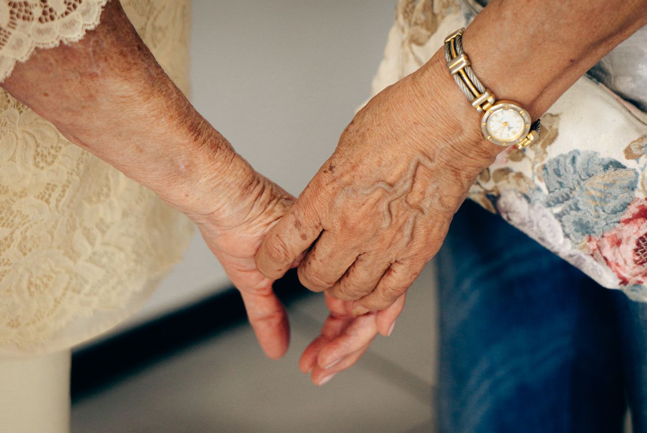 a younger and older woman holding hands, representing a daughter wanting to care for her elderly parents including through finding full-time specialized support