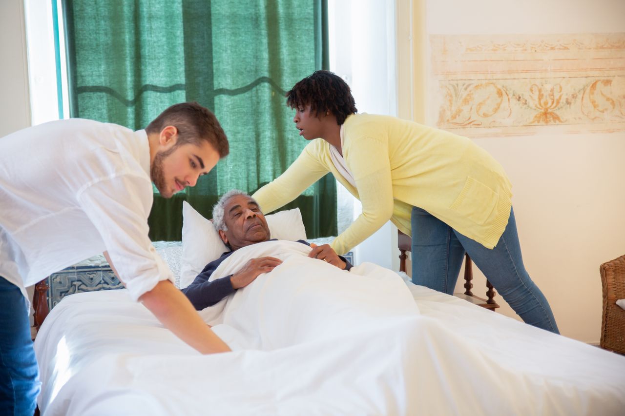 older man being tucked into bed by his loving daughter and son-in-law