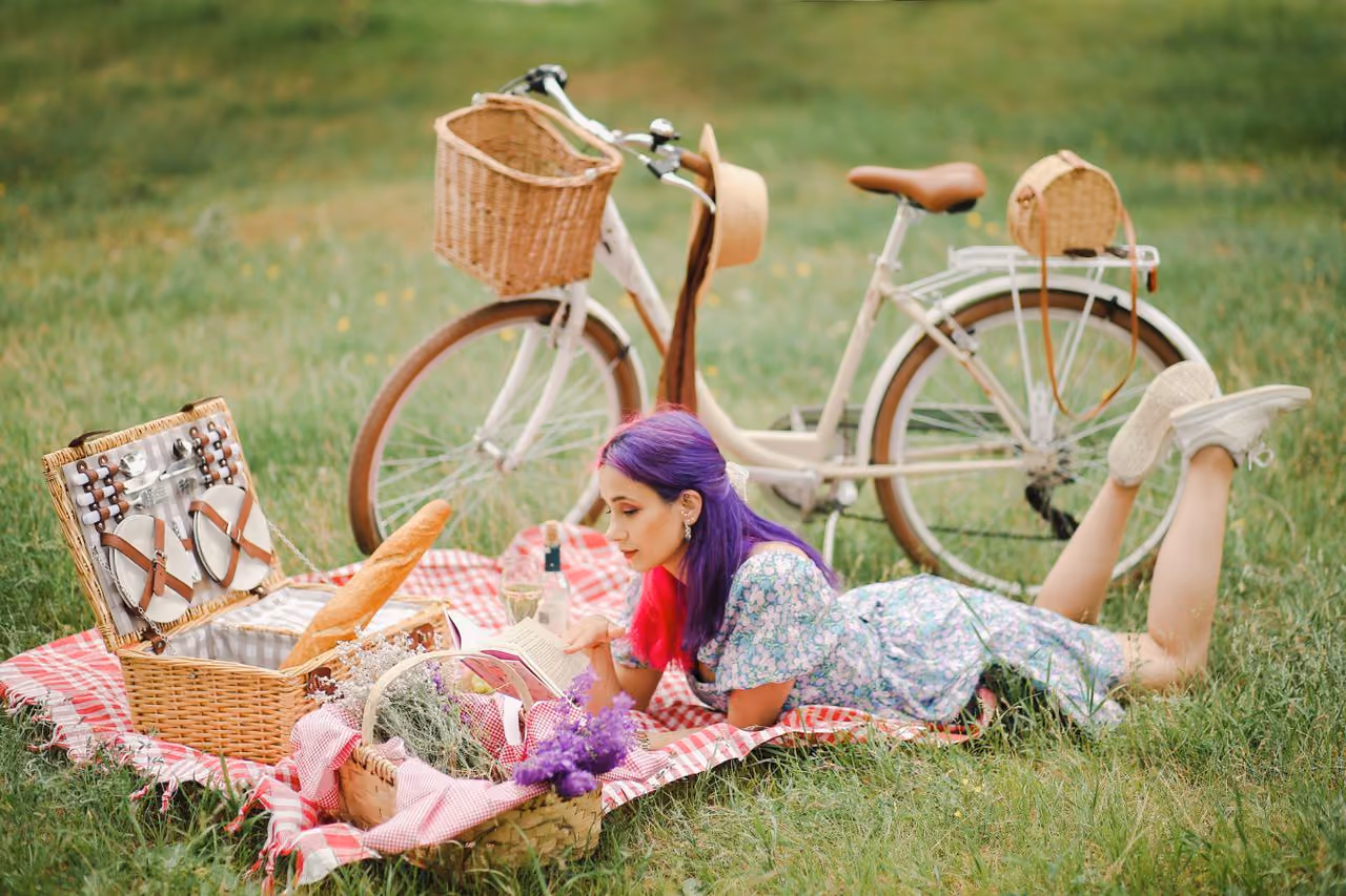 young woman with seemingly conflicting aesthetics - vibrant purple hair, a classic picnic, an old bike, reading a book in a field