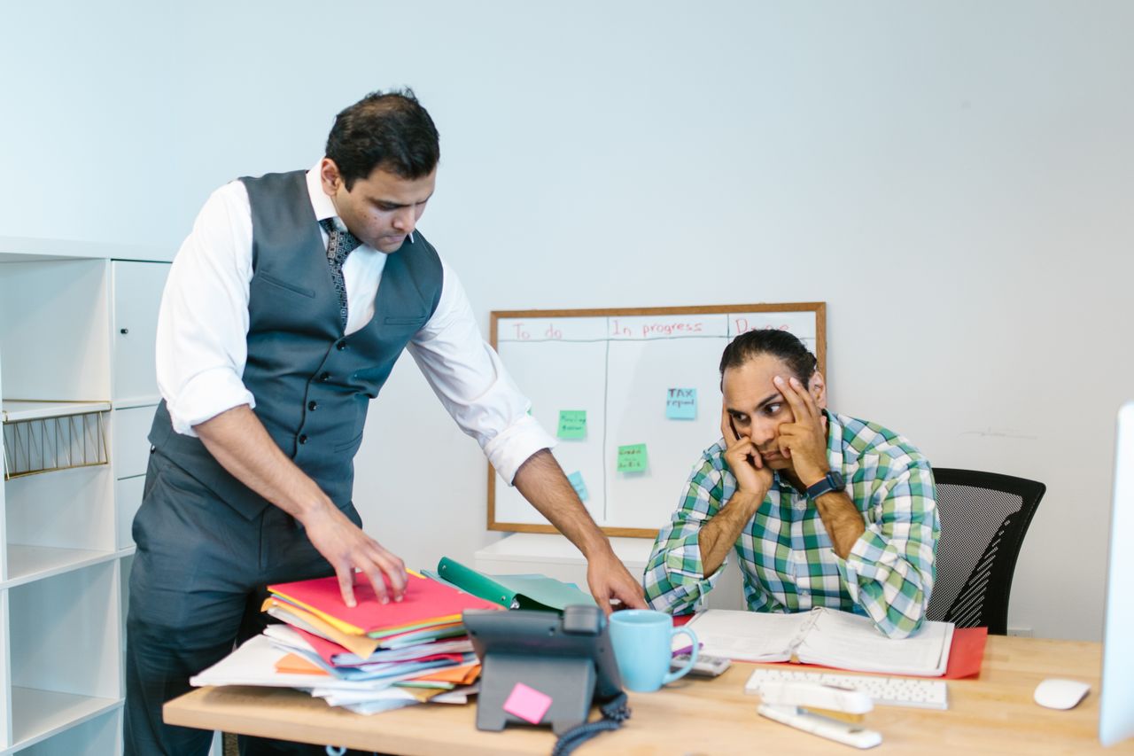 young professional sitting at a cluttered desk looking overwhelmed as his boss drops a stack of new folders in front of him