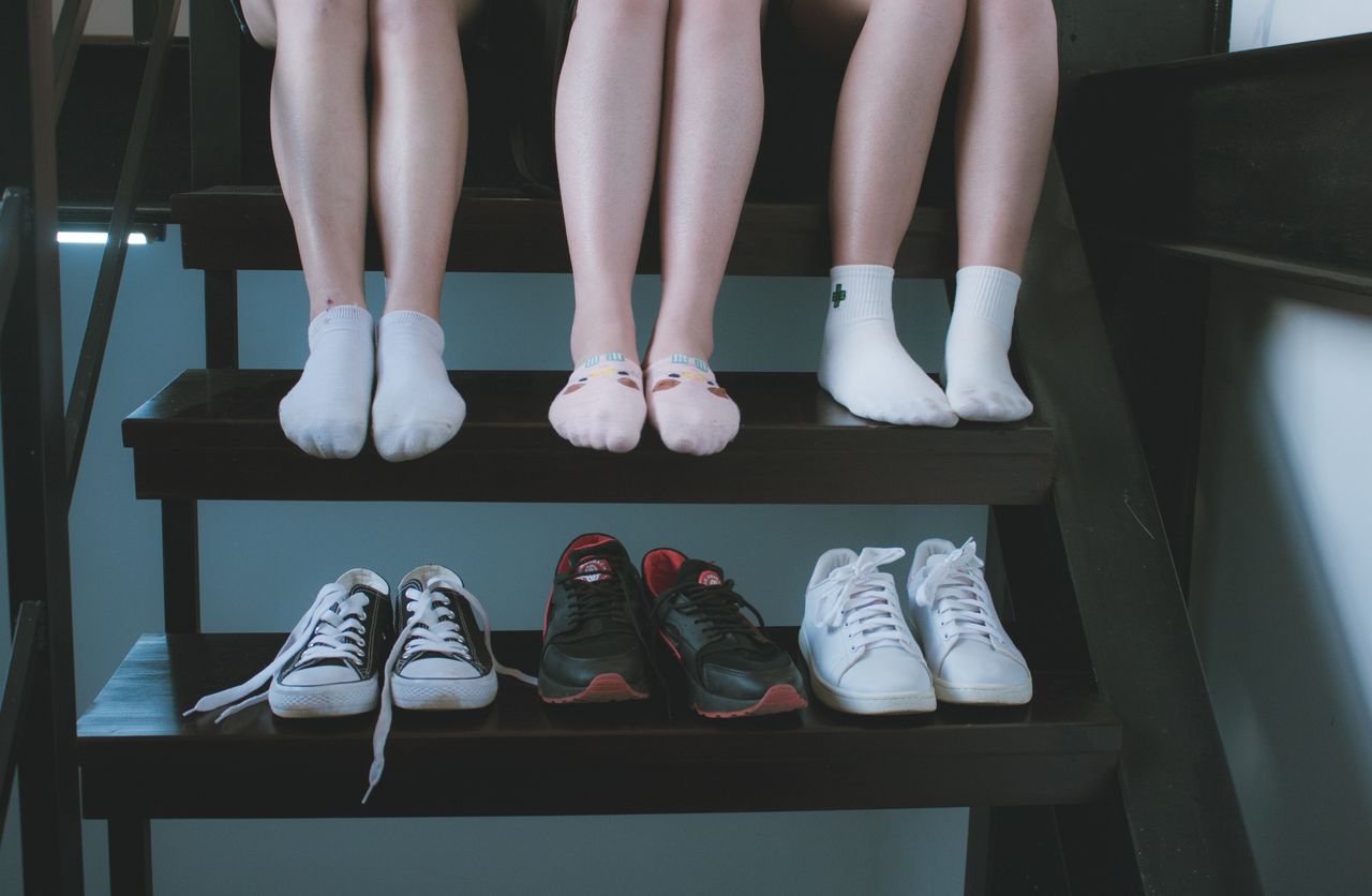 three women seated on a staircase in socked feet wearing socks like bombas with their shoes sitting on the stair beneath them