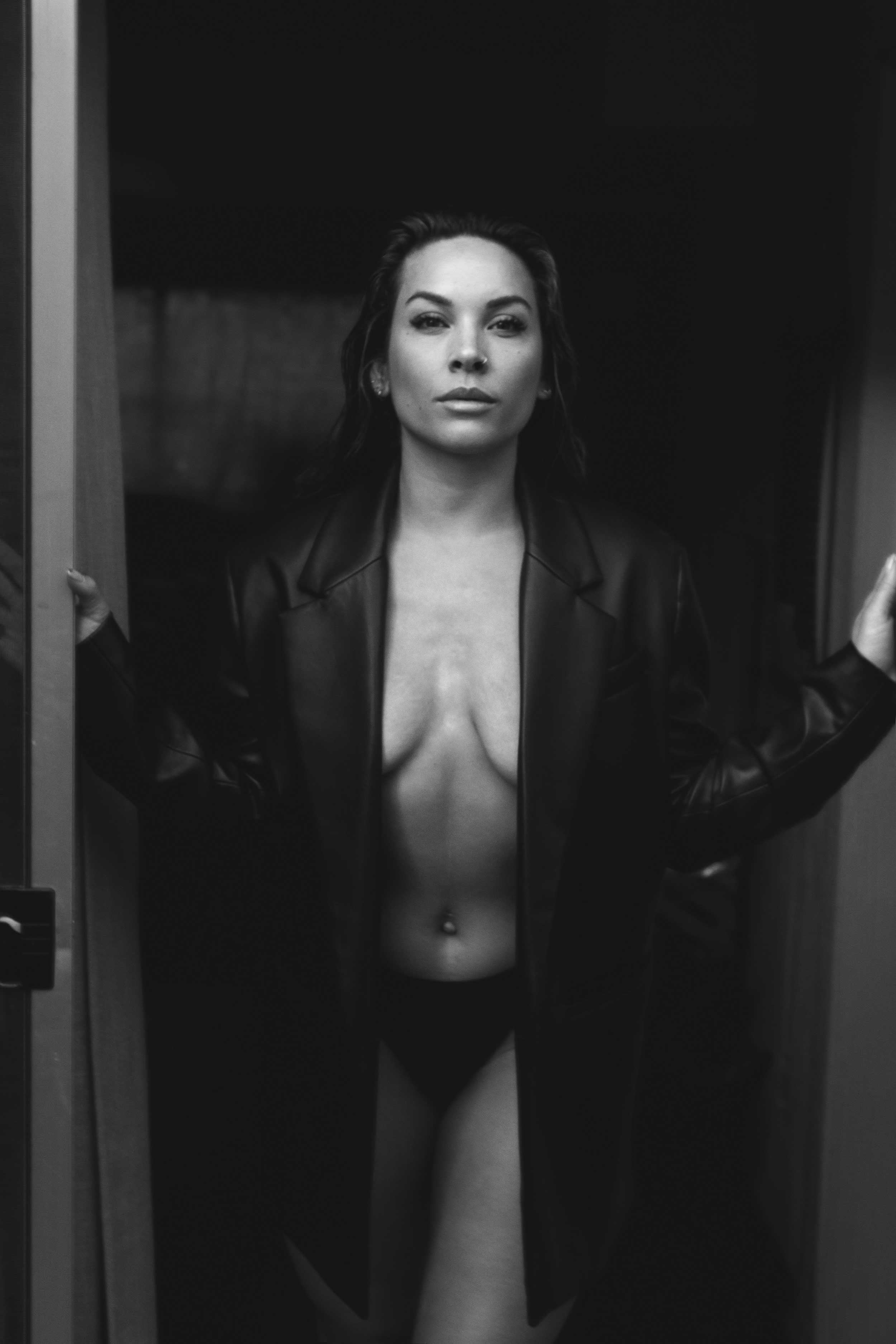 Black and white image of Apolonia Calleja wearing an oversized black coat, confidently posing braless in a bold, artistic portrait