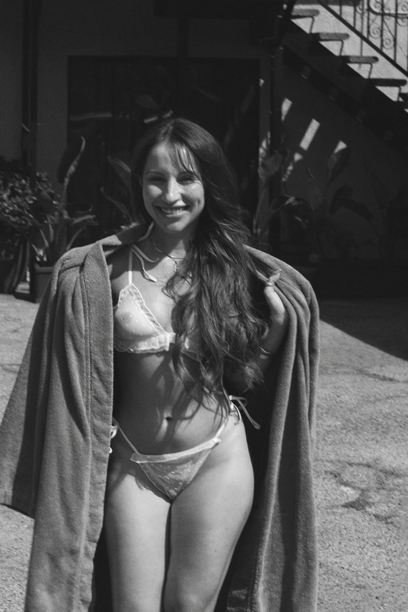 Arielle Lopez laughing joyfully outdoors in white vintage lingerie, holding a robe in the sunlight