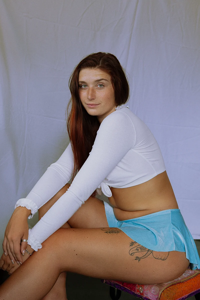 Color portrait of Britney Lee seated, red hair undone, makeup-free with visible tummy rolls, celebrating effortless beauty.