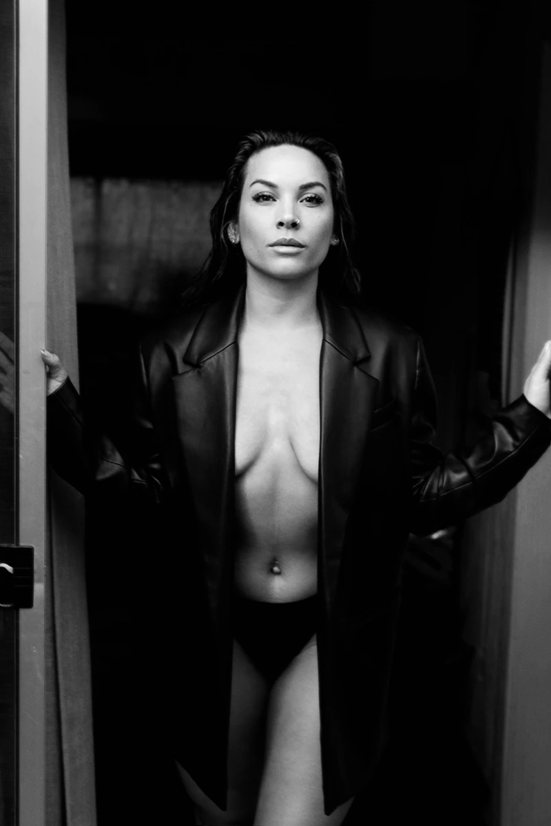 Black and white image of Apolonia Calleja wearing an oversized black coat, confidently posing braless in a bold, artistic portrait