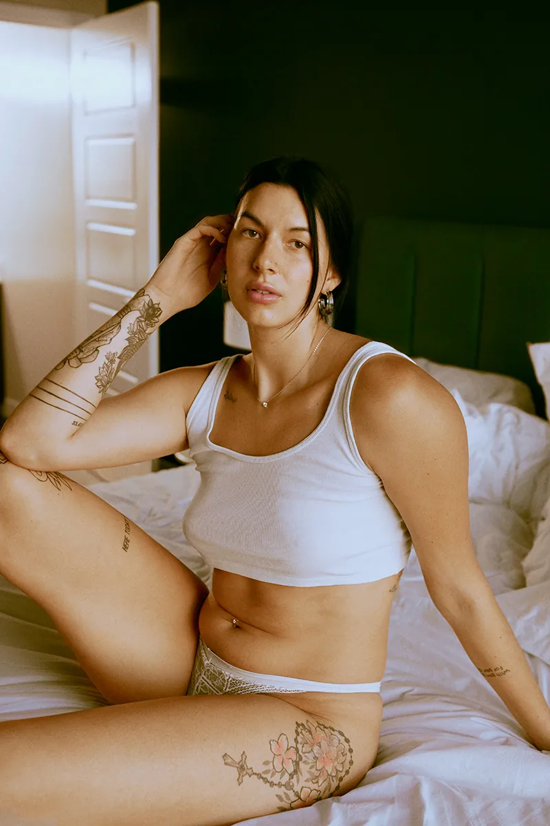 Willow Johnson sitting on bed in white tank top and underwear, no makeup, natural beauty in color photo