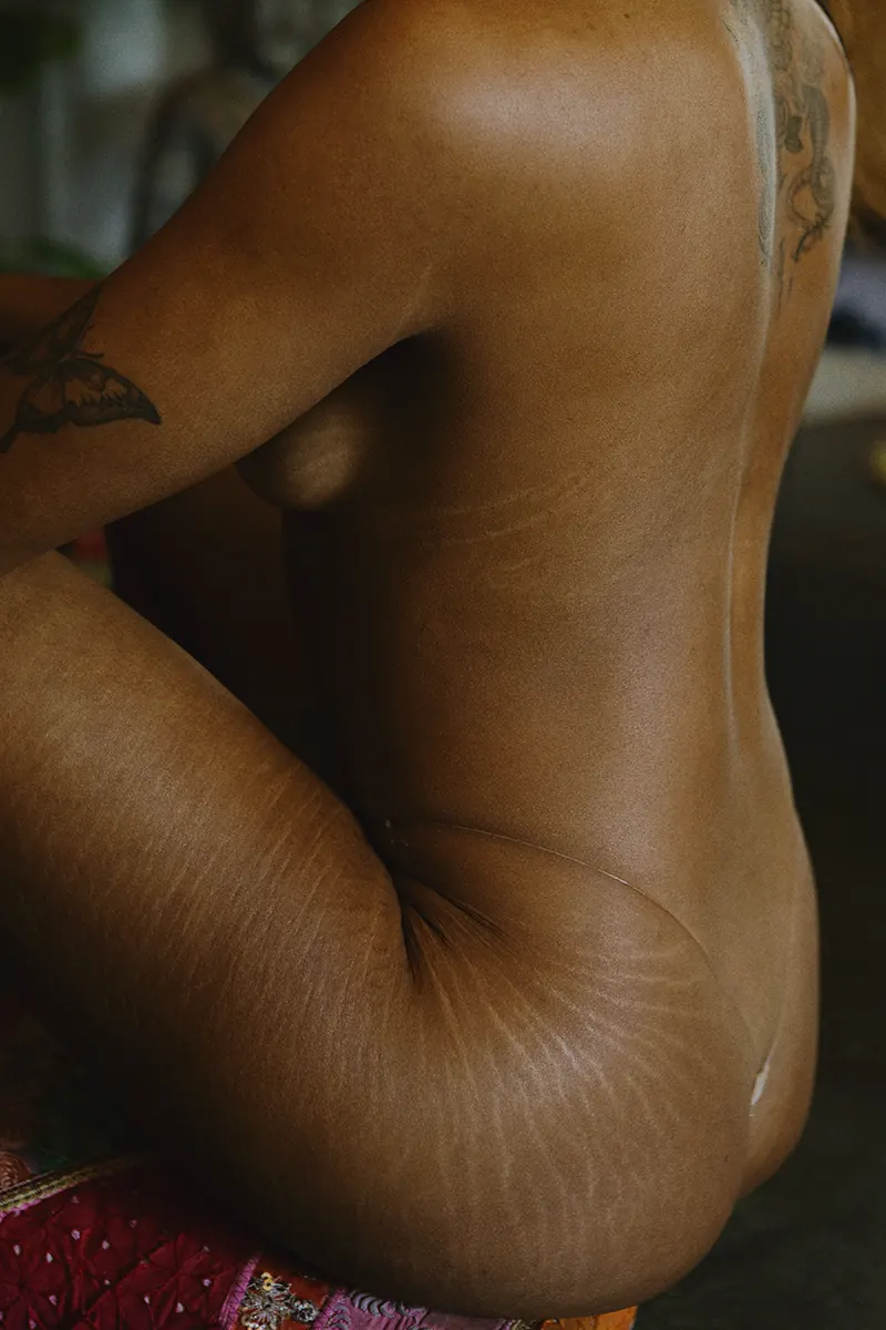 Color photo of a woman with stretch marks sitting on a stool, showing her back tattoo, glowing melanin, and natural beauty.