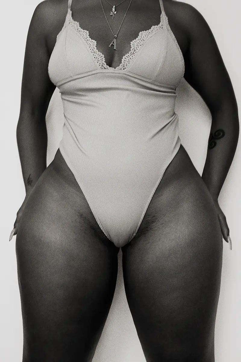 Black and white artistic photo of a Black woman standing in a one-piece swimsuit, natural beauty with thick thighs, body hair and ingrown hair visible, celebrating self-love and confidence.