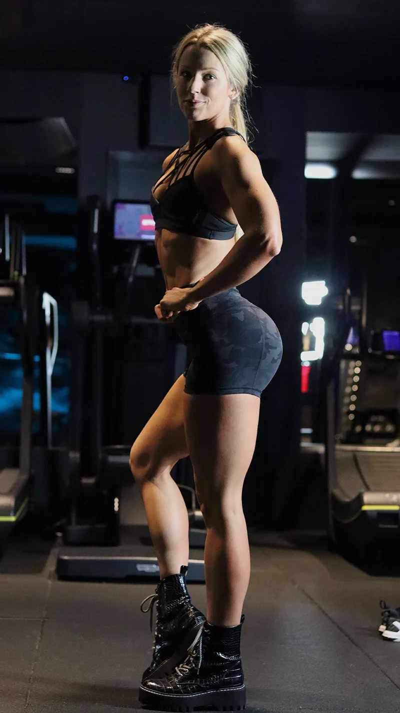 Fit woman in black sports bra and camo shorts posing confidently in the gym, highlighting toned glutes and strong quads.