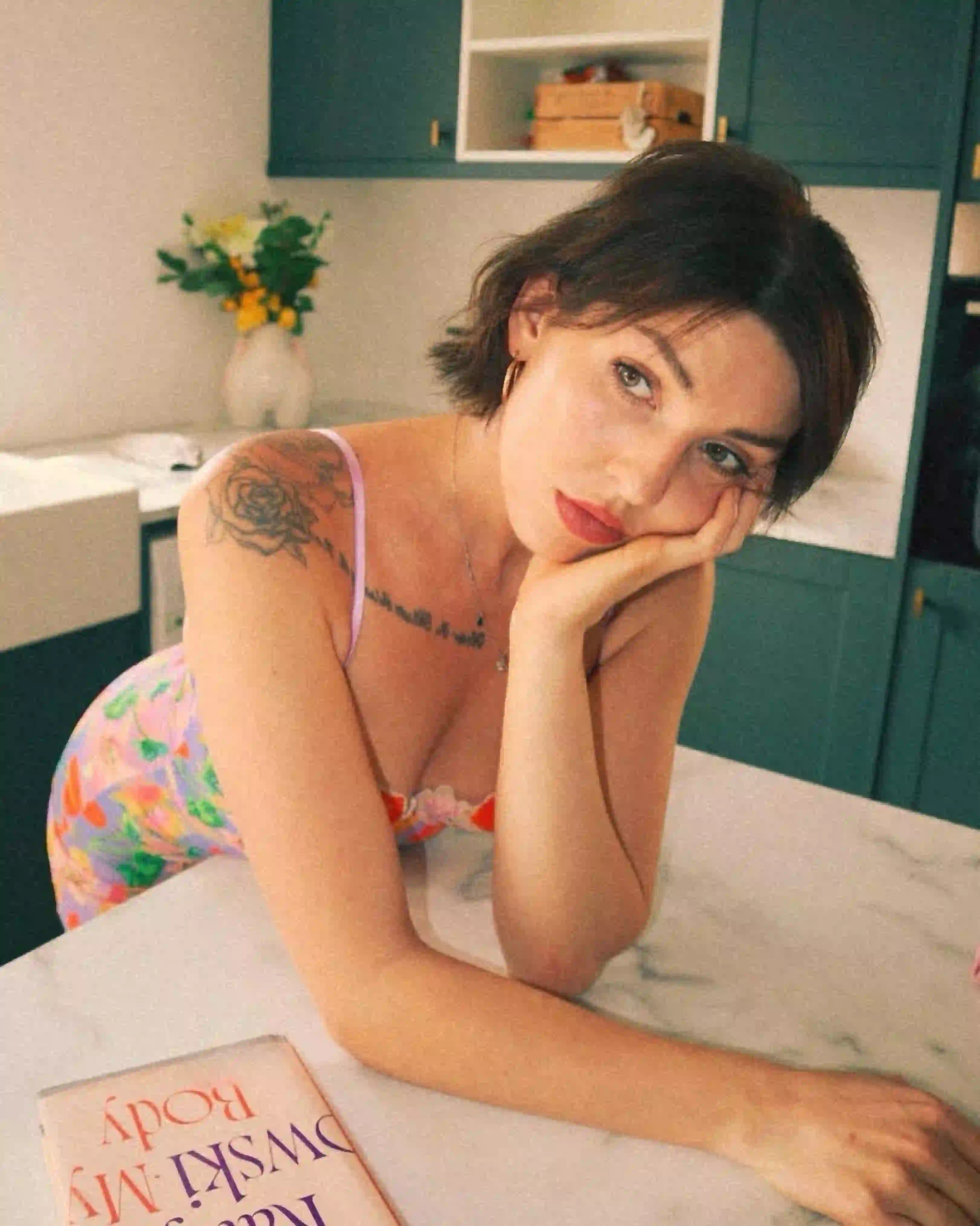 Woman resting her chin on her hand beside a book titled How to Dismantle My Body, reflecting on elf-love and body acceptance.