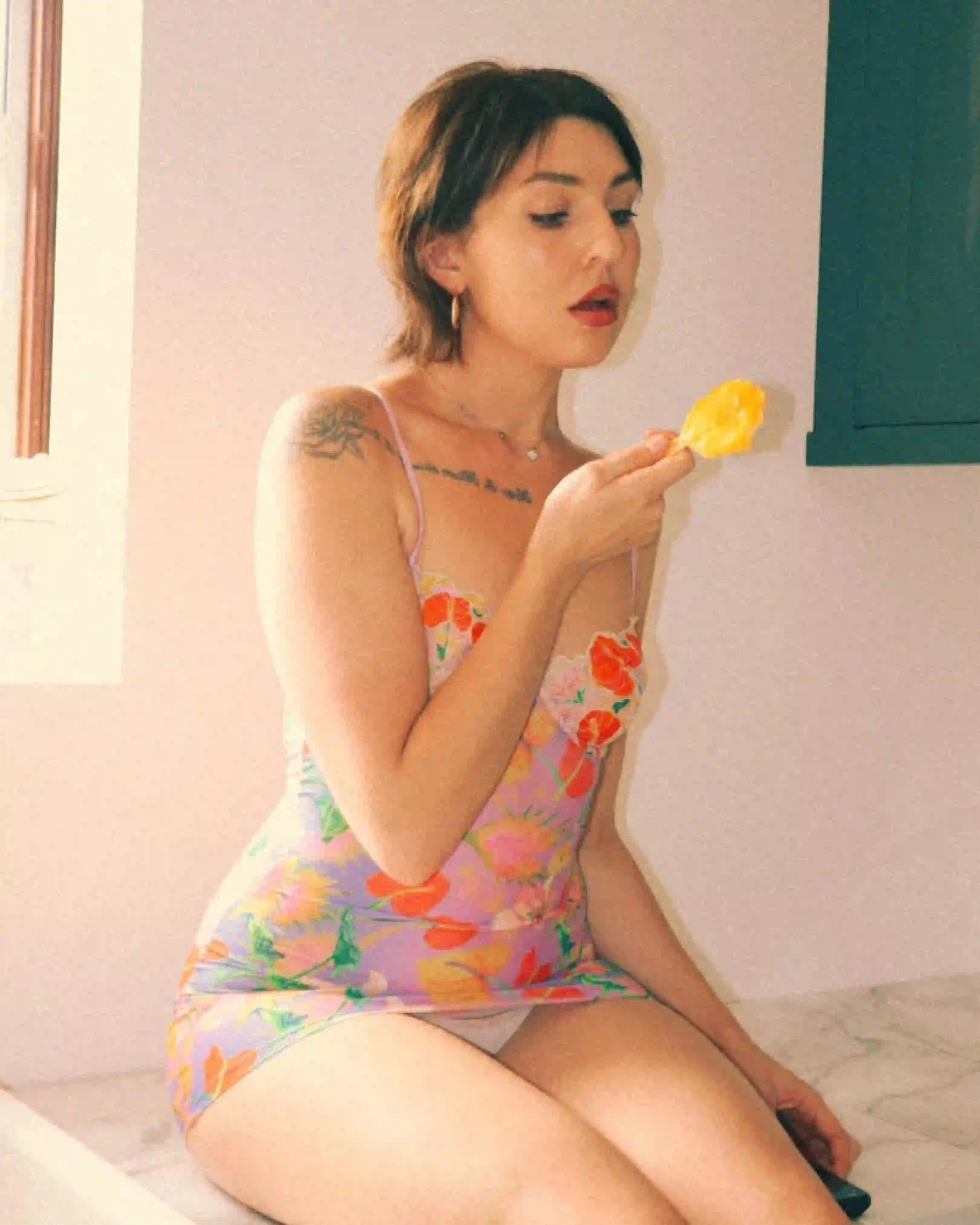 Woman in a floral lingerie slip sitting by the window, enjoying a popsicle with soft natural light and relaxed, confident energy.