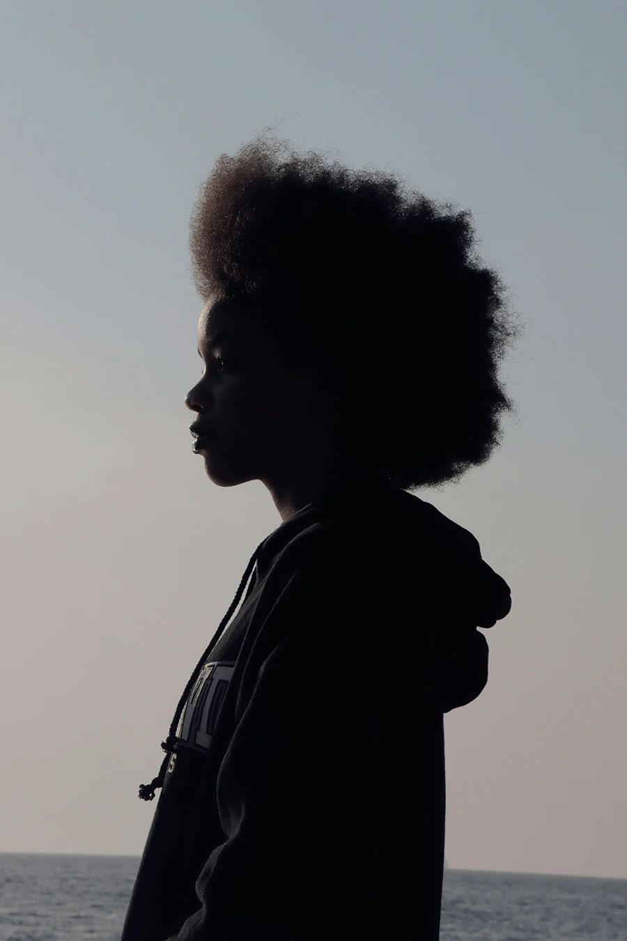 Black woman silhouette with natural afro standing near ocean at sunset, profile portrait.