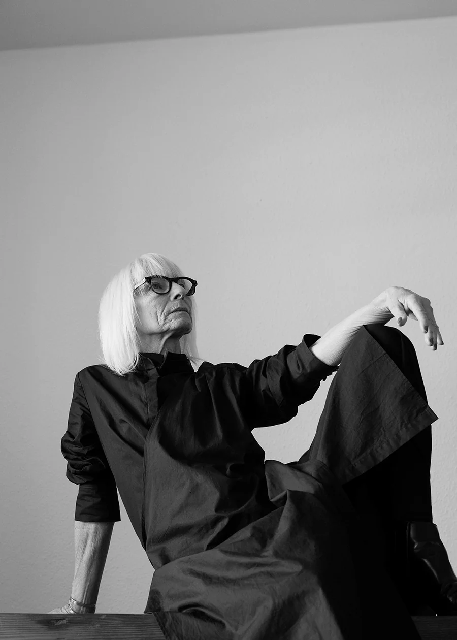 Black-and-white portrait of an older woman in minimalist fashion, seated in a sculptural pose.