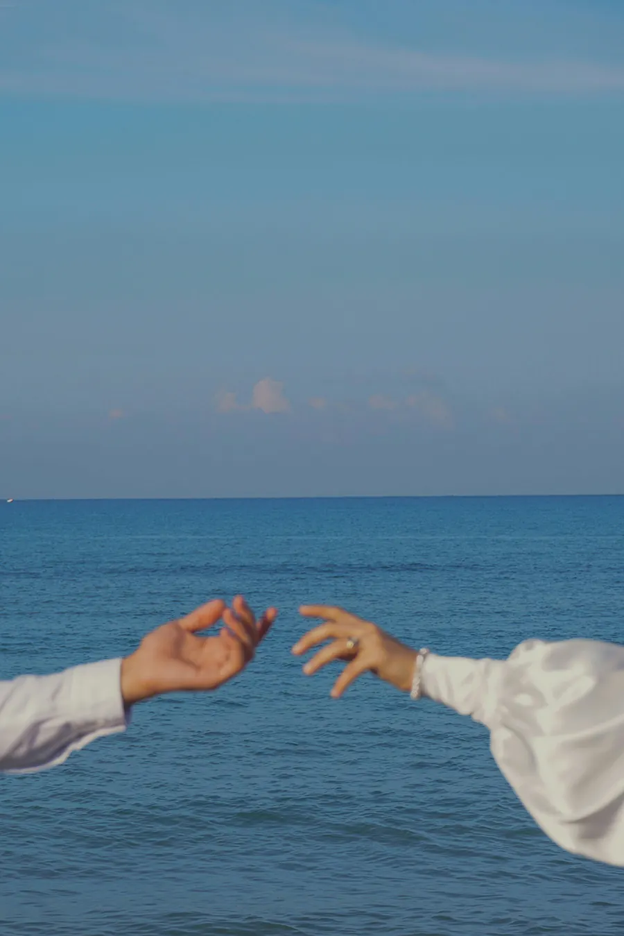 Two hands reaching toward each other above the ocean, sleeves in frame, with blue water and sky stretching into the horizon.