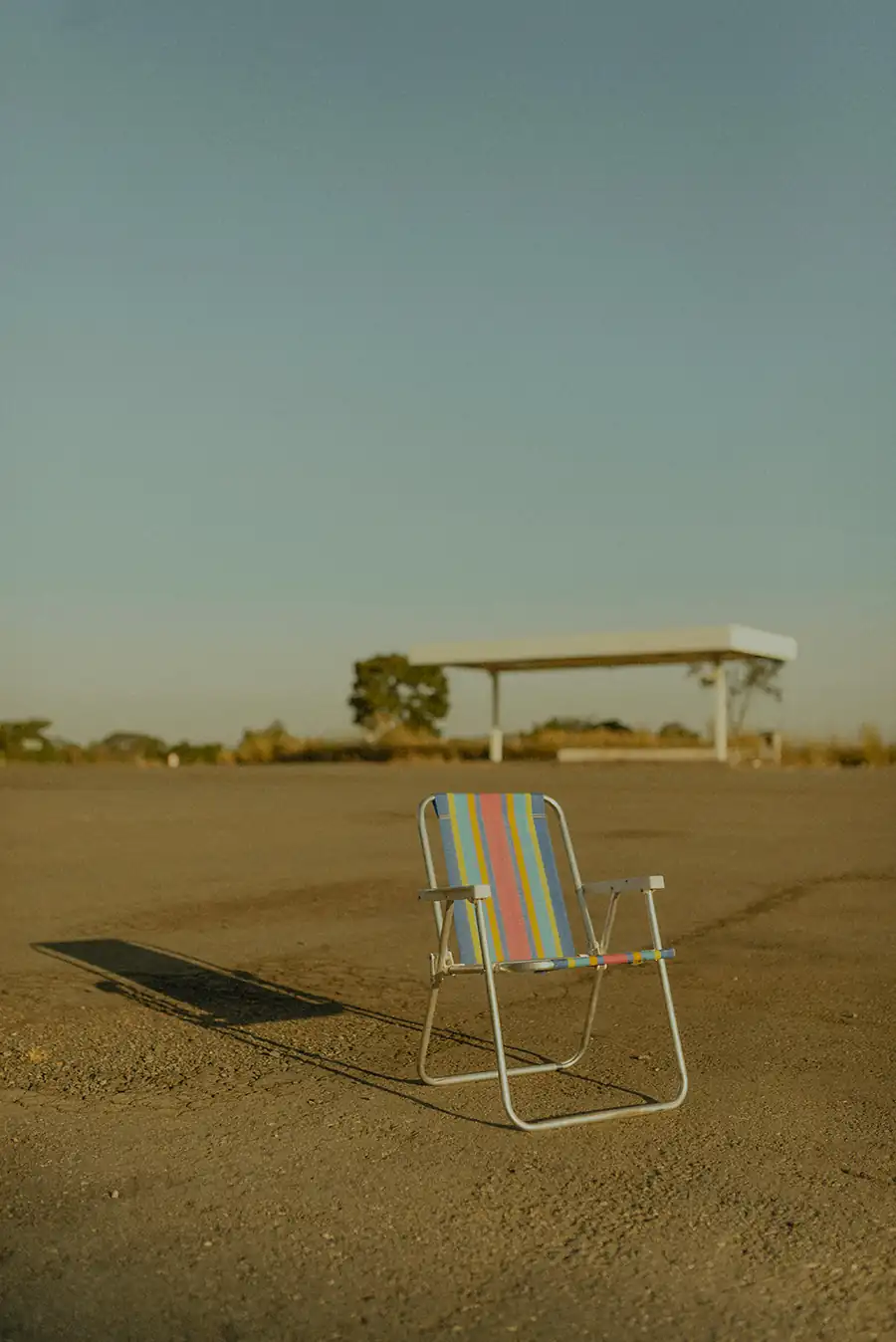 A single striped folding chair sitting alone in an empty sunlit open lot with long shadow and a distant gas station under a clear sky.