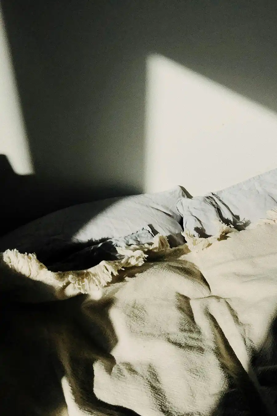 Unmade bed with sunlight and deep shadows on a pillow and blanket symbolizing restless sleep and anxiety.