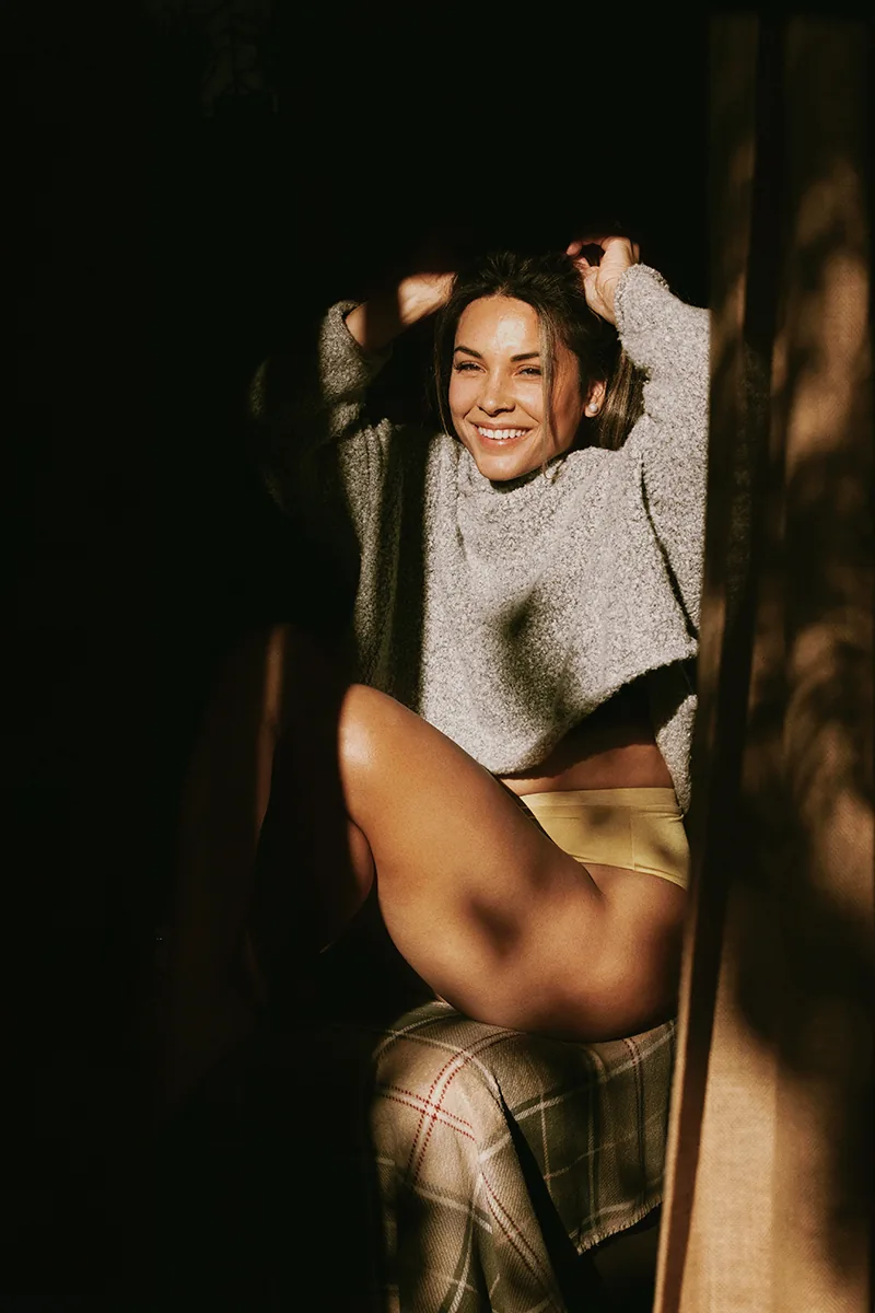 A smiling woman sitting in warm sunlight wearing a cozy sweater and underwear, legs highlighted by natural shadows and soft golden light.