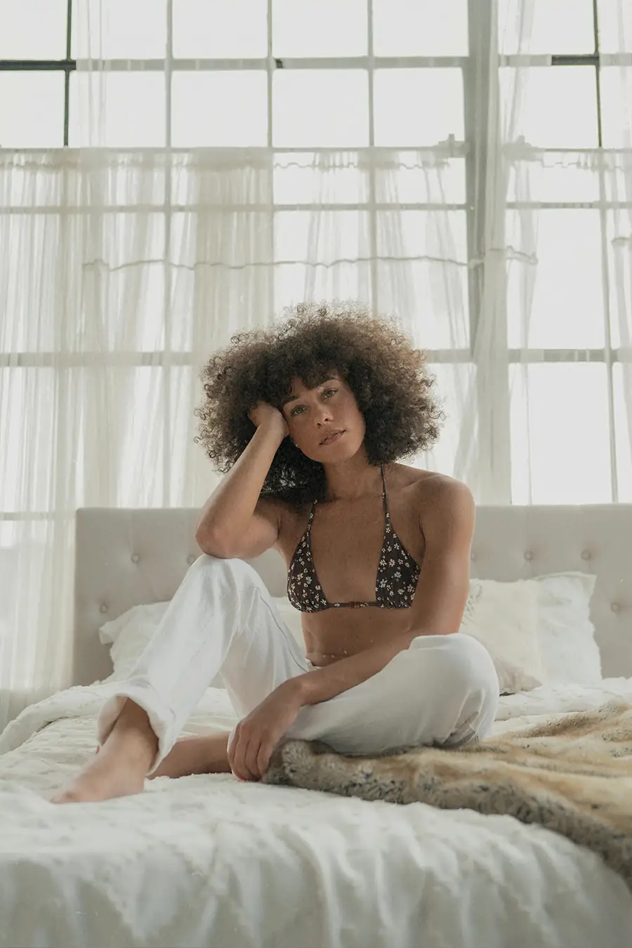 Woman sitting on a bed in soft morning light with large sheer curtains and industrial windows behind her.