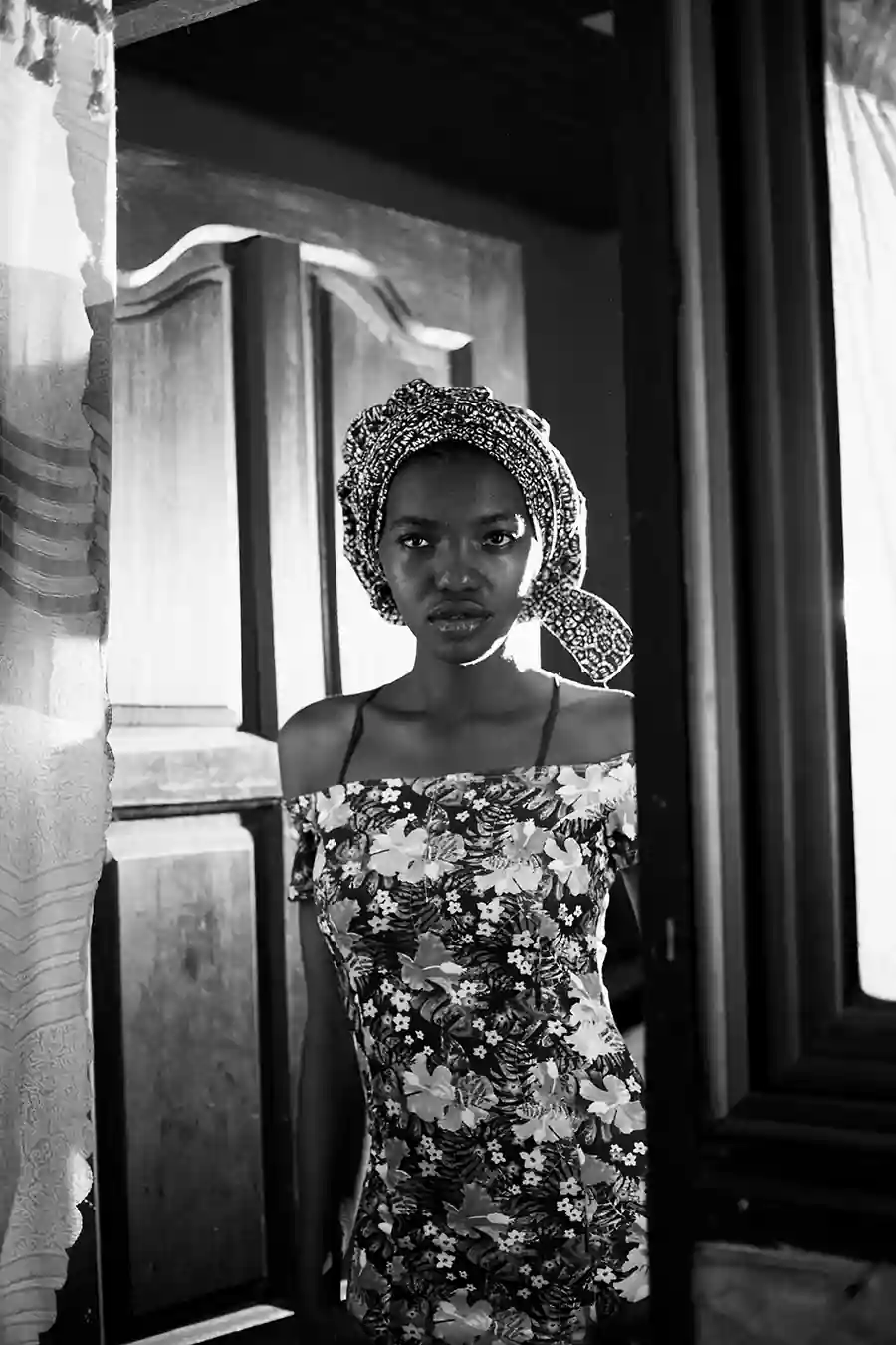 Black woman standing in a doorway wearing a floral dress and headwrap, looking directly at the camera in natural light. Portrait capturing identity, cultural expression, and the lived experience of how Black women are seen.