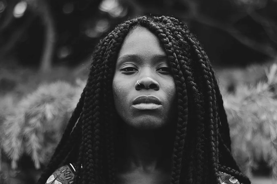 Black woman with long braids looking directly at the camera in a black-and-white close-up portrait, representing identity, natural hair, and the lived experience of being seen and judged.
