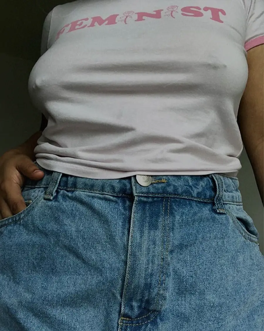 Close-up of a woman wearing a pink feminist T-shirt and high-waisted blue jeans, with a hand resting in the pocket.