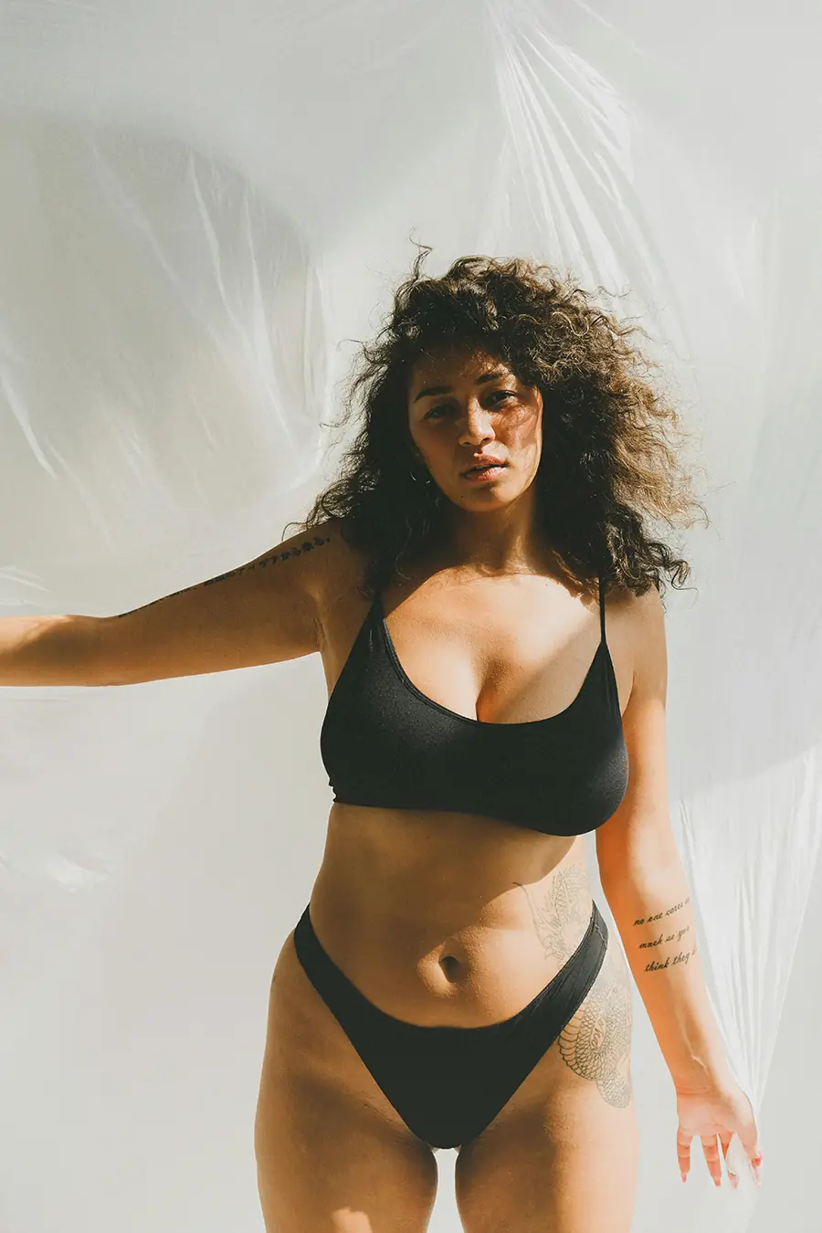 Woman standing in natural sunlight wearing a black bra and underwear, showing her curves, skin texture, and tattoos against a white backdrop.