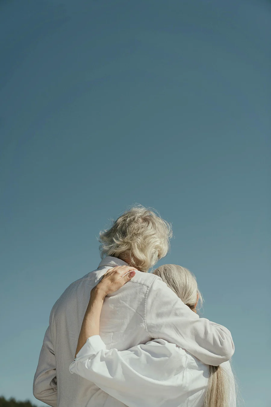 “Elderly couple embracing outdoors with their backs to the camera under a clear blue sky, symbolizing support, connection, and lifelong companionship.
