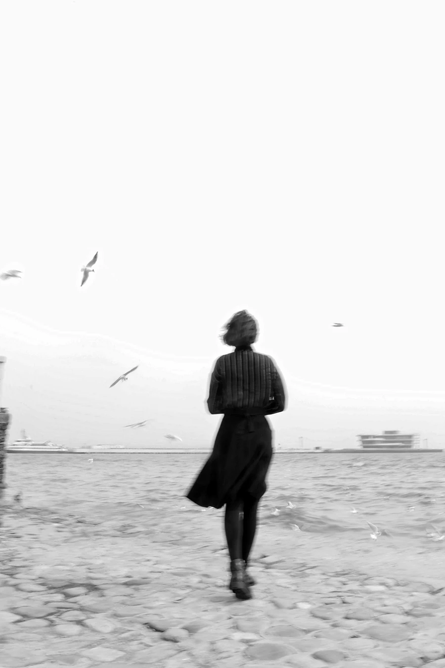 Woman walking alone on a stone path by the ocean, her silhouette softened by motion blur as seagulls circle above in a quiet, reflective scene captured in black and white.