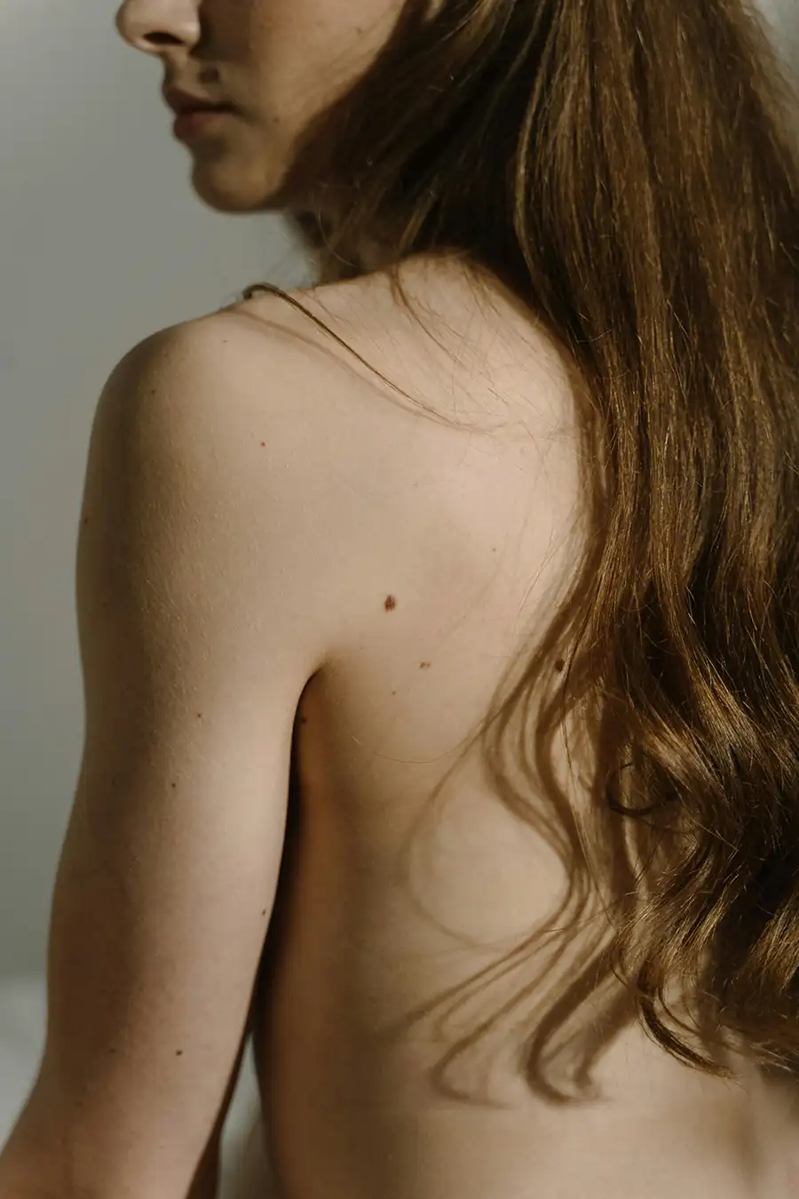 A soft, intimate portrait of a woman with long hair and bare shoulders turned in profile, captured in natural light with a quiet, introspective mood.